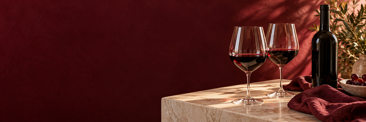 Two glasses of red wine on a warm ivory table against a deep burgundy background